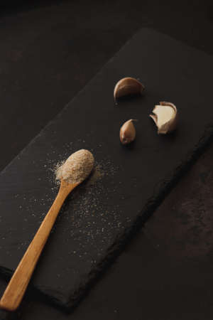 Composition of garlic powder sprinkled into wooden spoon and thre cloves on black slate board. Copyspace. Selective focusの写真素材
