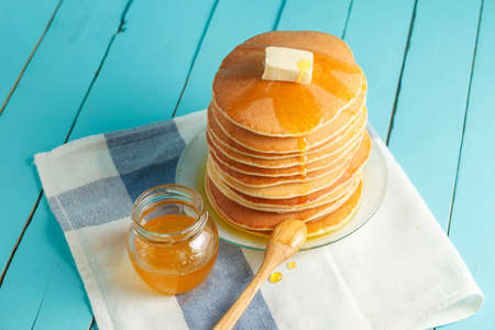 Stack of pancakes with pouring honey on plate with wooden spoon and jar with honey. Concept of tasty...