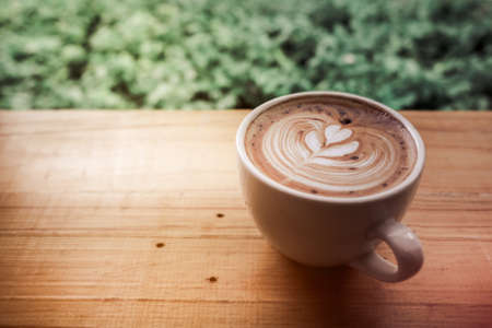 A cup of coffee latte art with heart in a white cup with green leave frame on wooden backgroundの写真素材