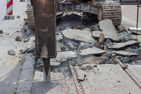 Tram track reconstruction and excavator with hydraulic hammer breaking asphalt. Demolition, ground works, and works on the superstructures in the reconstruction of tram tracks. Selective focus.の写真素材
