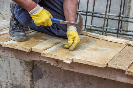 Builder hands hammering a nail into a plank, formwork installation at construction site. Selective...