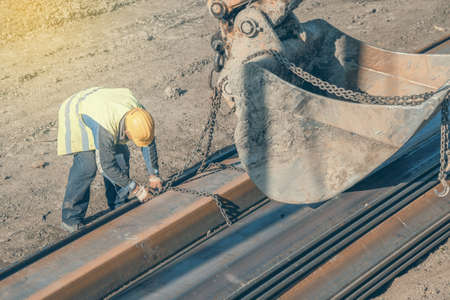 Construction worker attaching lifting chains on steel profile for excavator bucket lifting. Made...