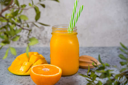 Mango smoothie with orange in a jar and with tubes stands on a gray concrete background, green leaves around. Clean and healthy food for vegetarians. Healthy diet.の写真素材