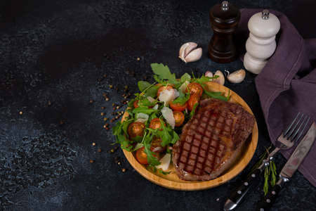 A juicy roast beef steak with fresh salad in a wooden plate stands to the right of the frame on a black concrete background. Horizontal orientation with copy space.の写真素材