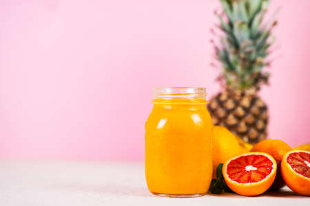 A small jar of yellow vegan smoothie stands on a pink background, surrounded by oranges and fresh...