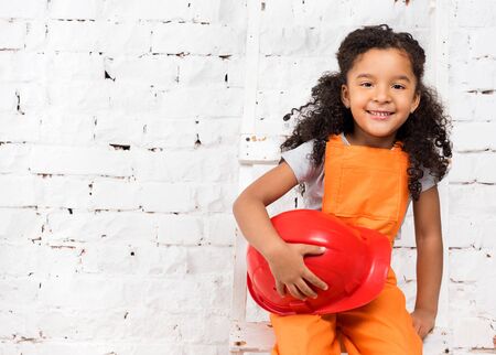pretty little girl in repairman uniform with helmet in hands sitting on ladderの写真素材