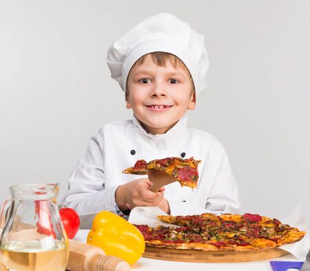funny little boy in cook uniform holding a piece of tasty pizzaの写真素材
