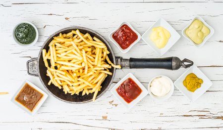 French fries in frying pan with different sauces in white saucers on white wooden background with cracked paint. Top view. View from above.の写真素材