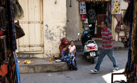 Zanzibar, Tanzania - July 15, 2016: Muslim woman feeding her little child on the street of zanzibar, man walking neaby, dirtyのeditorial素材