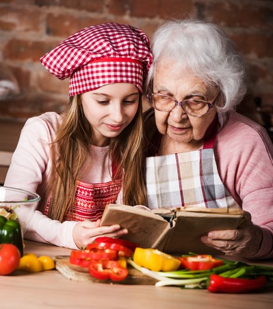 Little granddaughter reading recipe book with her granny sitting at kitchenの写真素材