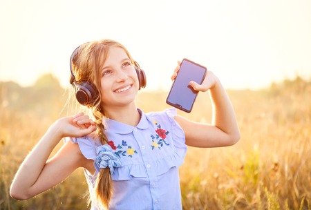 Smiling teenage girl hold smartphone and listening to music with joyfully raised hands in the field