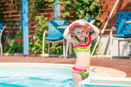 Little smiling blond girl in a bright swimsuit holding her hat standing in the blue poolの写真素材