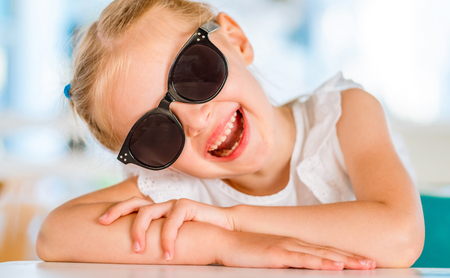 Little smiling blond girl in black sunglasses sitting at the table and inclined her headの写真素材