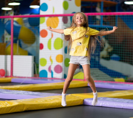 Little excited girl in a yellow T shirt jumping on the trampoline on the colorful amusement park background