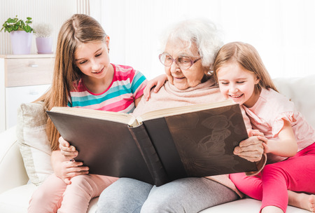 Grandmother with little smiling granddaughters reading big brown book togetherの写真素材