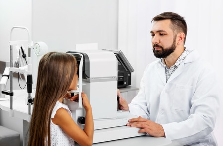 Little blond girl undergoes eye survey on special modern diagnostics device with doctor in the ophthalmologic clinicの写真素材