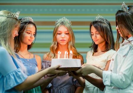 Pretty smiling teenage girls in dresses and crowns holding cake at birthday party od