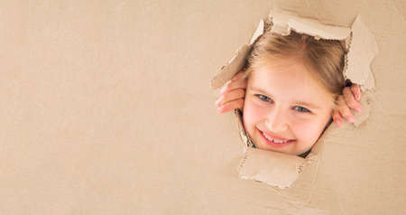 Funnt little girl looking through the torn hole in cardboard boxの写真素材