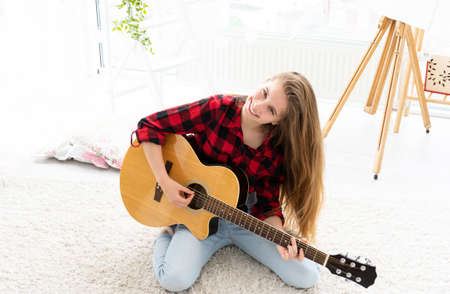 Cute girl with long loose hair playing guitar in light roomの写真素材