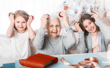 Smiling granddaughters holding decorative balls like eyes with grandma at Christmas