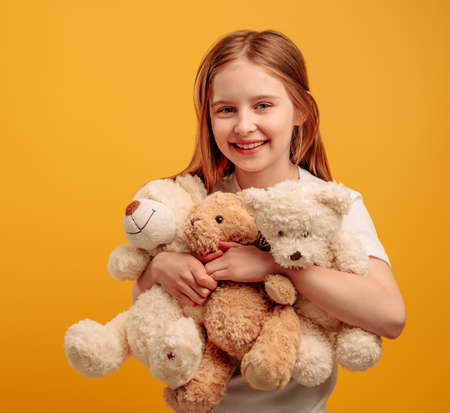 Beautiful girl child with three teddy bears isolated on yellow background with copyspace. Portrait of kid holding toys and smiling looking at the cameraの写真素材