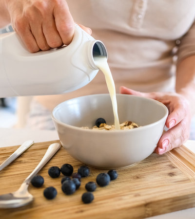 Girl puts milk to oatmeal with blueberry in bowl plate for delicious cereal breakfast at kitchen. Woman preparing granola with berry and latte for morning healthy diet mealの写真素材