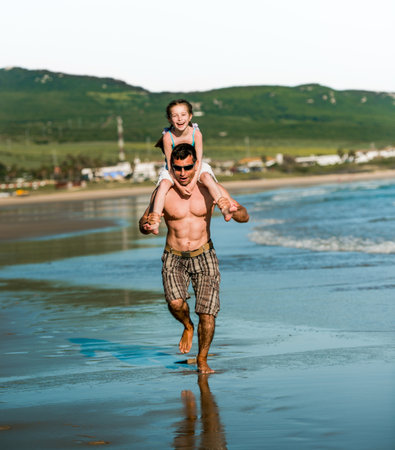 Father holding daughter at shoulders at sea coast and running. Family dad and pretty smiling child kid at beach at summerの写真素材