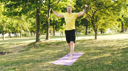 Athletic young man doing an exercises for the shoulders with the elastic rubber band on a grass...