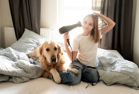 Cute Little Girl Drying Hair With A Hairdryer, Sitting With Dog On A Bedの写真素材