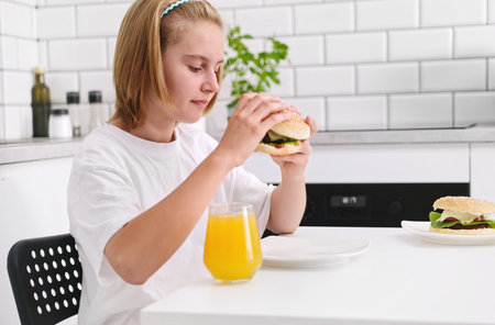 Cute Little Girl Eating A Fresh Meat Burger In The Kitchenの写真素材