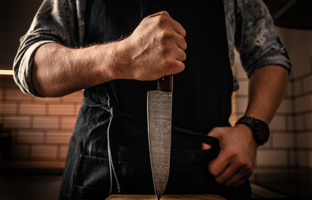 Chief man in apron holding knife vertically at kitchen stubbing wooden board. Restaurant proffesional with culinary tool closeupの写真素材