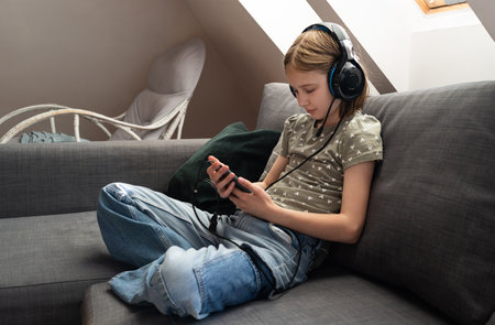 Cute Little Girl Browsing Social Media On Smartphone, Wearing Headphones On Sofa At Home