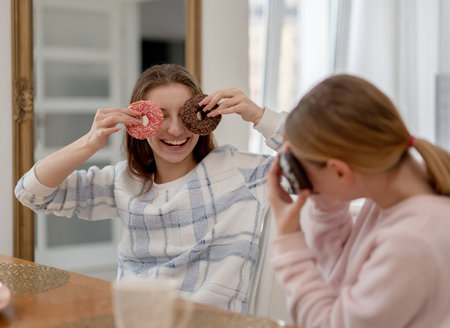 Two Smiling Sisters In Pajamas Have Fun Playing With Donuts At Breakfast