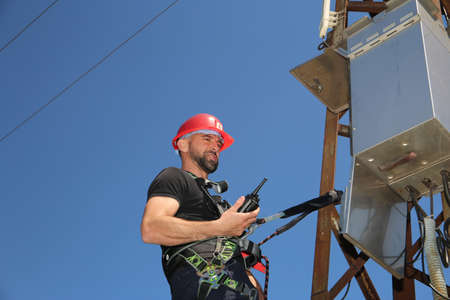 Tehnician in red helmet and safety belt with radio station on the electric pole