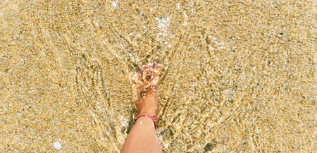 The leg of a girl entering the clear sea water on the beach of a tropical island. Yellow sand and glittering streams of water in the sunの写真素材