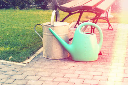 Rubber and tin watering cans near a bench in the summer garden. Sunny morning raysの写真素材