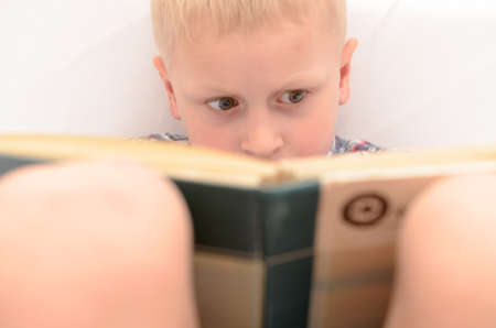 A five-year-old boy is reading a book on the bed.の写真素材