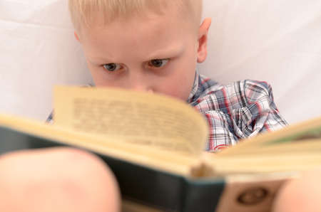 A five-year-old boy is reading a book on the bed.の写真素材