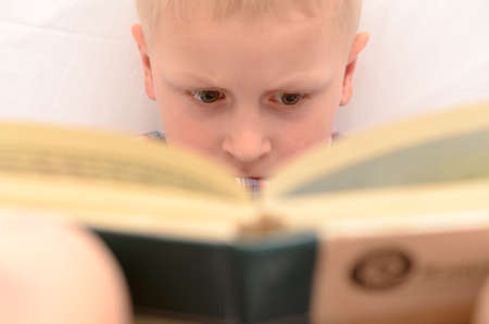 A five-year-old boy is reading a book on the bed.の写真素材
