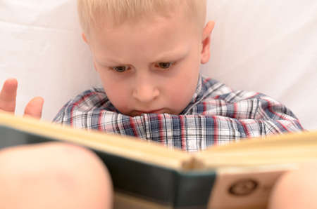 A five-year-old boy is reading a book on the bed.の写真素材