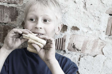 A hungry dirty boy greedily eats a crust of bread against the wall