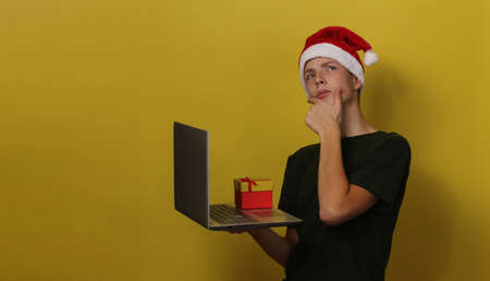 n Santa Claus hat and red t-shirt holding gray laptop and red box. eenage boy in Santa hat wearing...