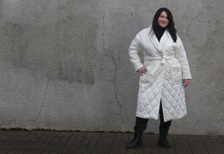 Portrait of a woman plus size model with dark hair in a white XXL winter coat and black trousers posing against wall.の写真素材