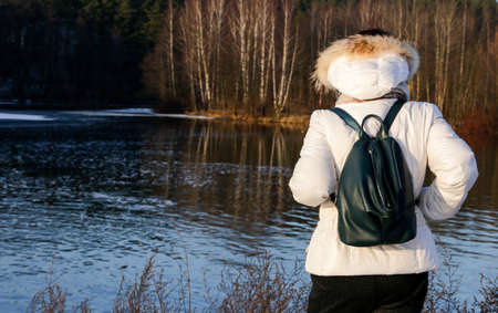 Woman in white jacket with dark backpack standing on the river bank with her back to the camera.の写真素材