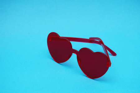 Red sunglasses in the shape of a heart on a blue background. Elegant red plastic glasses in the shape of a heart on a blue paper background close-up.の写真素材