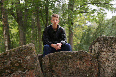 A handsome young man in a blue hiking jacket from the rain sits on a large stone in the background of a forest with trees. Boy tourist on vacation.の写真素材