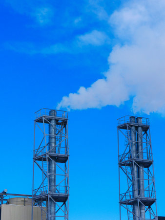 Two chimneys of a heating plant against the background of a bright blue sky and smoke coming out of them.の写真素材
