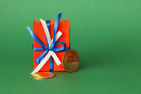 A red gift box with a blue and white ribbon and two yellow bitcoin coins stands on a green paper background.....の写真素材