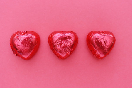 Three heart-shaped chocolates wrapped in red foil on a red background, top view.