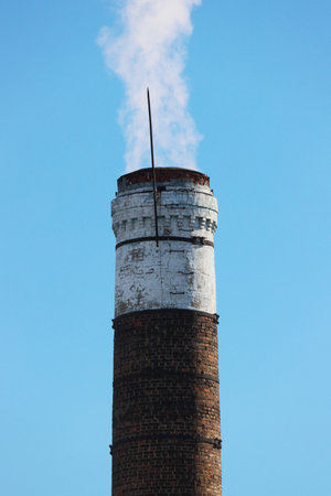 An old smoking brick chimney of a power plant against the background of a blue, clear sky. Factory smoke causes pollution in the air. Fuming chimney against blue sky.の写真素材
